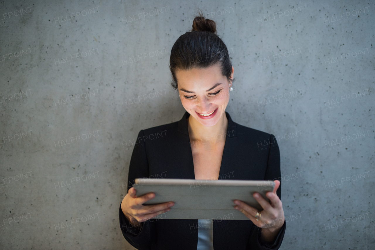 A portrait of happy young business woman with digital tablet standing against concrete wall in office.