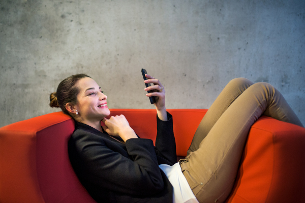 A young businesswoman with smartphone sitting on red armchair in office, a gray concrete wall in the background.