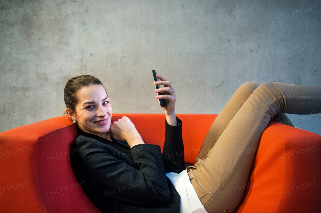 A young businesswoman with smartphone sitting on red armchair in office, a gray concrete wall in the background.