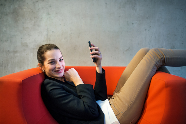 A young businesswoman with smartphone sitting on red armchair in office, a gray concrete wall in the background.