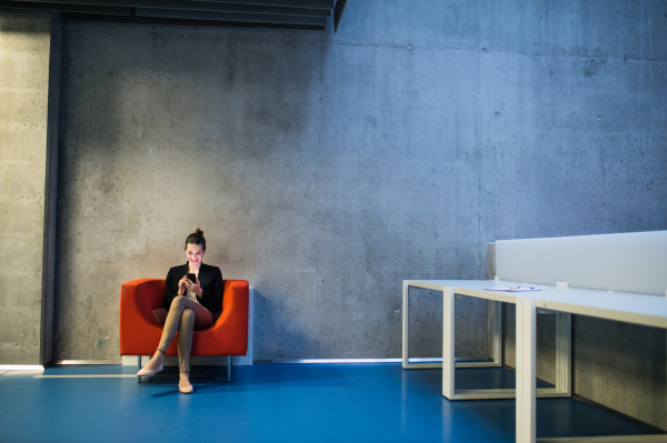 A young businesswoman with smartphone sitting on red armchair in office, a gray concrete wall in the background. Copy space.