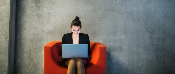 A young businesswoman with laptop sitting on red armchair in office, a gray concrete wall in the background. Copy space.