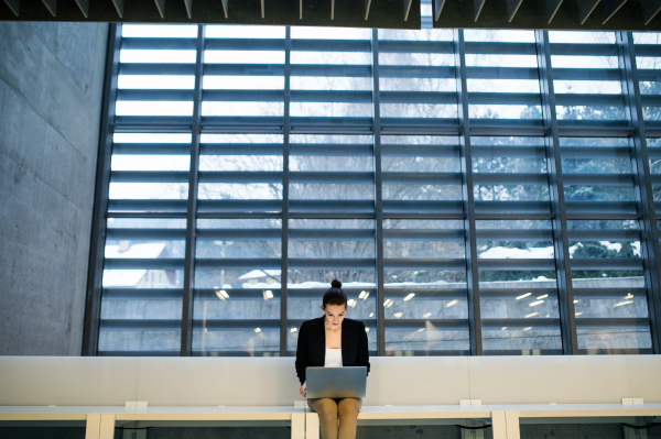 A portrait of a young student or businesswoman sitting on desk in room in a library or office, using laptop.