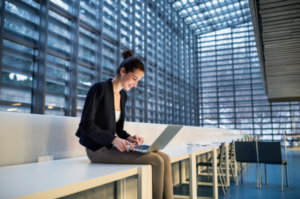 A portrait of a young student or businesswoman sitting on desk in room in a library or office, using laptop.