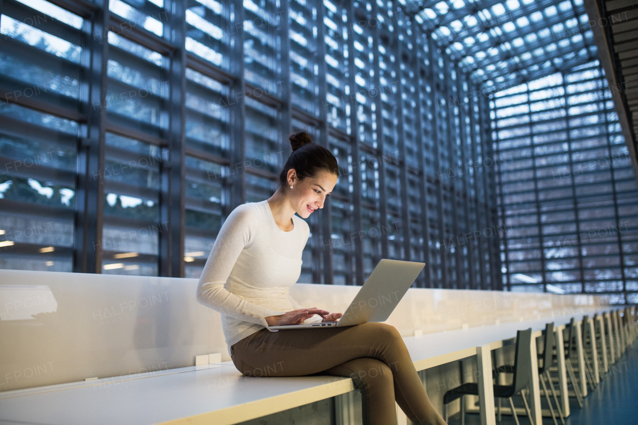 A portrait of a young student or businesswoman sitting on desk in room in a library or office, using laptop.