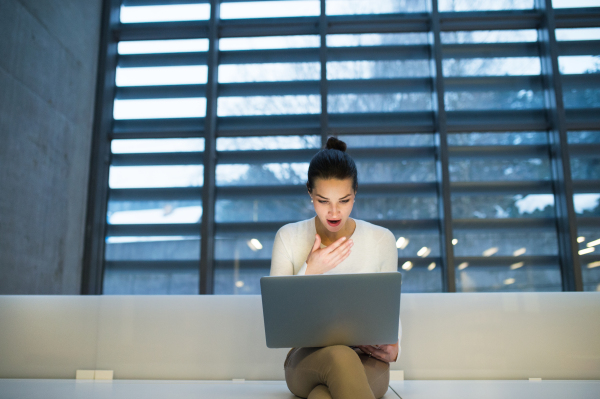 A portrait of a young student or businesswoman sitting on desk in room in a library or office, using laptop. Copy space.