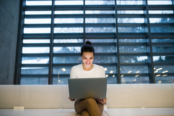 A portrait of a young student or businesswoman sitting on desk in room in a library or office, using laptop.