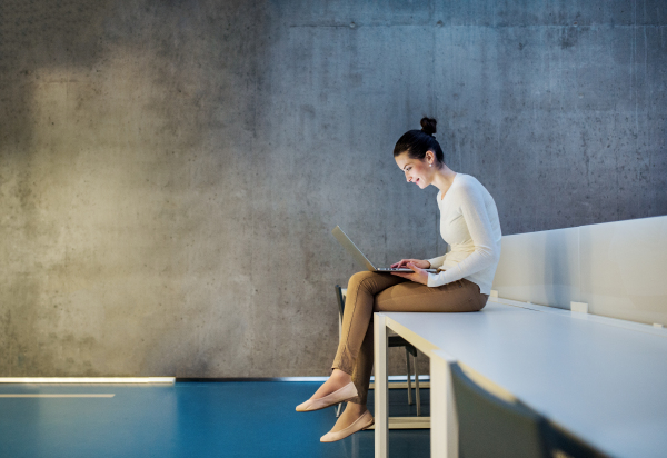 A portrait of a young student or businesswoman sitting on desk in room in a library or office, using laptop.