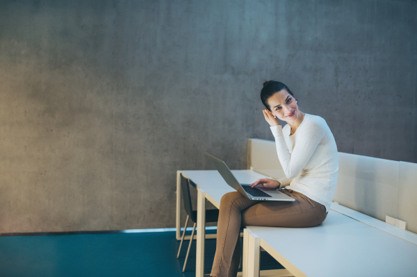 A portrait of a young student or businesswoman sitting on desk in room in a library or office, using laptop. Copy space.