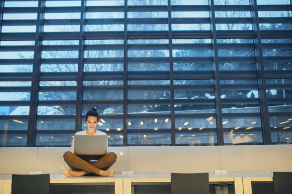 A portrait of a young student or businesswoman sitting on desk in room in a library or office, using laptop. Copy space.