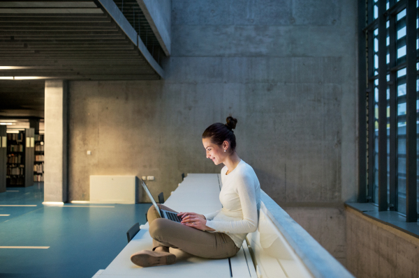 A portrait of a young student or businesswoman sitting on desk in room in a library or office, using laptop.