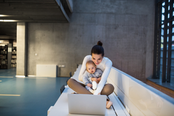 A portrait of a young student mother or businesswoman sitting on desk with a baby in room in a library or office, using laptop.