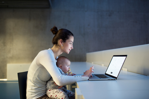 A side view of a young student mother or businesswoman sitting on desk with a baby in room in a library or office, using laptop.