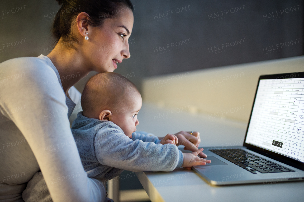 A side view of a young student mother or businesswoman sitting on desk with a baby in room in a library or office, using laptop.