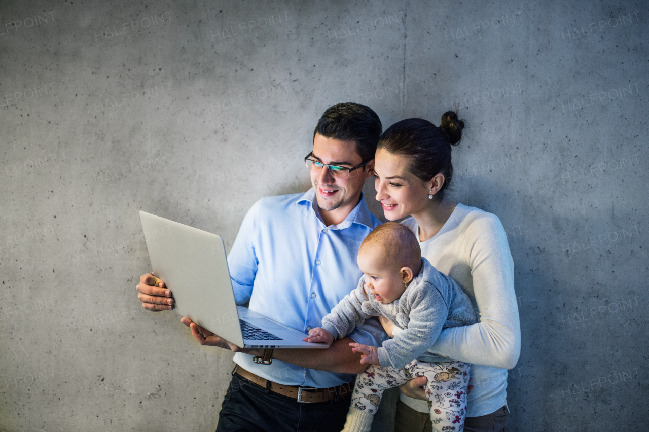 A young businessman with wife and baby daughter standing together in office, using laptop.