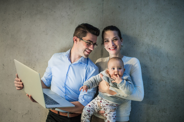 A young businessman with wife and baby daughter standing together in office, using laptop.