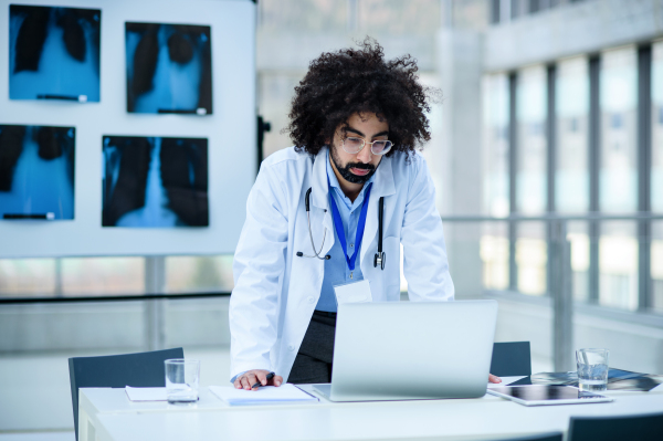 Front view portrait of serious male doctor standing in hospital, using laptop.