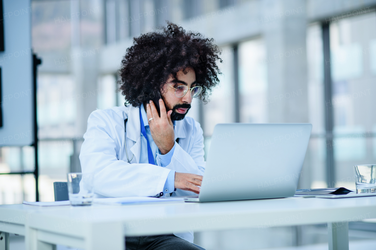 Portrait of busy male doctor sitting in hospital, using laptop and smartphone.