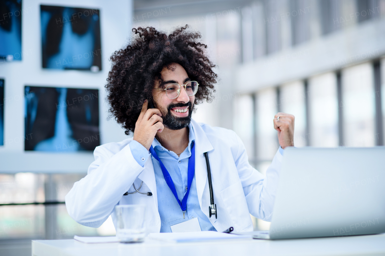 Portrait of cheerful male doctor sitting in hospital, using laptop and smartphone.