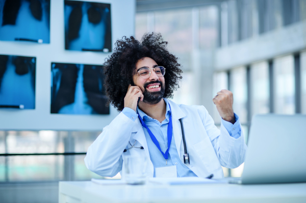 Portrait of cheerful male doctor sitting in hospital, using laptop and smartphone.