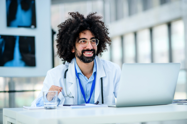 Portrait of cheerful and happy male doctor sitting in hospital, using laptop.