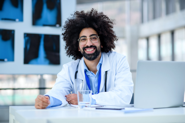 Front view portrait of cheerful male doctor sitting in hospital, looking at camera.