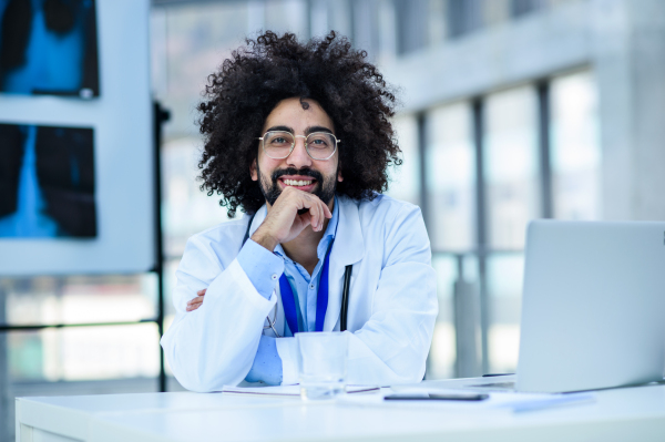 Front view portrait of cheerful male doctor sitting in hospital, looking at camera.