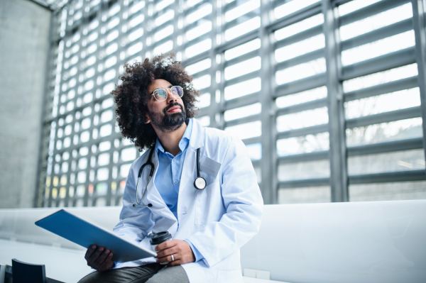 Portrait of thoughtful male doctor with clipboard sitting in hospital. Copy space.