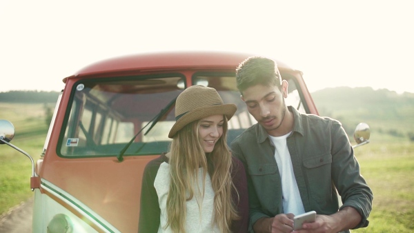 Young couple on a roadtrip through countryside standing by a minivan, using map on smartphone. Slow motion.