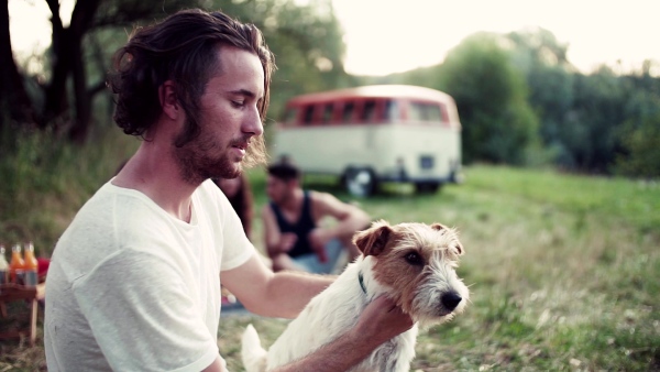 A young man with a dog and friends sitting on grass on a roadtrip through countryside. Slow motion.