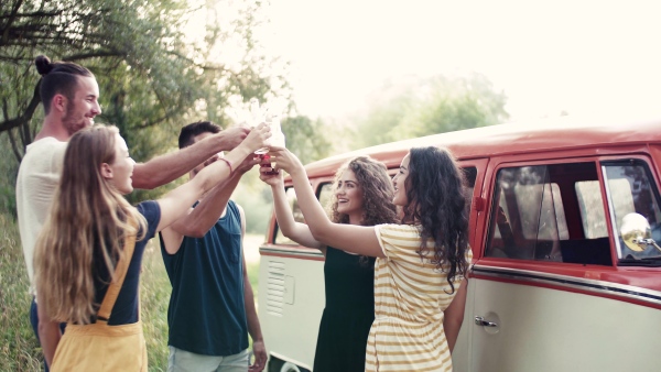 A group of young friends with drinks standing outdoors on a roadtrip through countryside, clinking bottles.