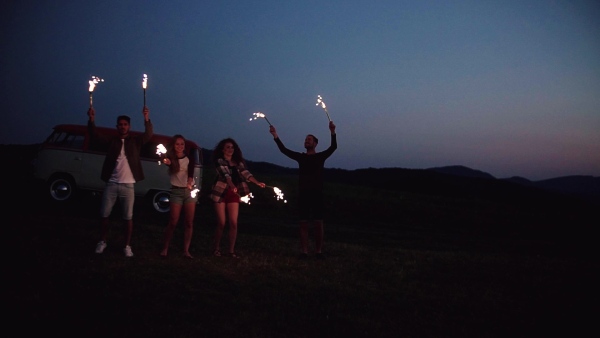 A group of young friends with sparklers dancing outdoors on a roadtrip through countryside at dusk. Slow motion.