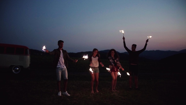 A group of young friends with sparklers standing outdoors on a roadtrip through countryside at dusk. Slow motion.