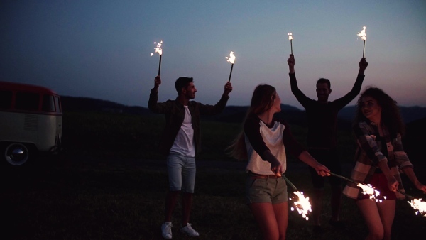 A group of young friends with sparklers standing outdoors on a roadtrip through countryside at dusk. Slow motion.