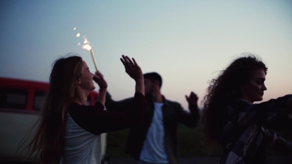 A group of young friends with sparklers dancing outdoors on a roadtrip through countryside at dusk. Slow motion.