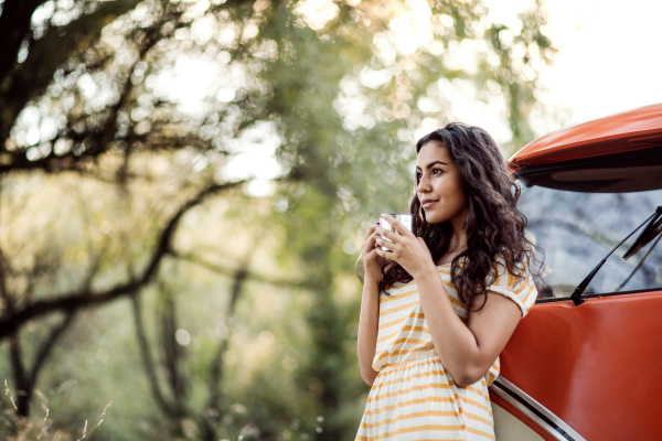 A cheerful young girl with coffee standing by a car on a roadtrip through countryside. Copy space.