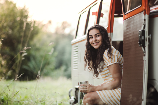 A cheerful young girl with coffee standing by a car on a roadtrip through countryside. Copy space.