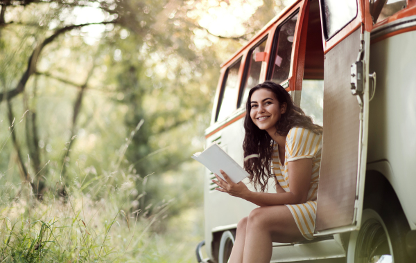 A cheerful young girl with book by a car on a roadtrip through countryside, reading. Copy space.