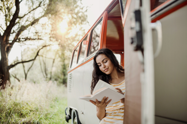 A cheerful young girl with book by a car on a roadtrip through countryside, reading. Copy space.