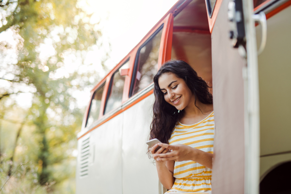 A cheerful young girl with smartphone by a car on a roadtrip through countryside, texting. Copy space.