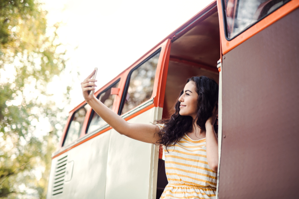 A cheerful young girl with smartphone by a car on a roadtrip through countryside, taking selfie.