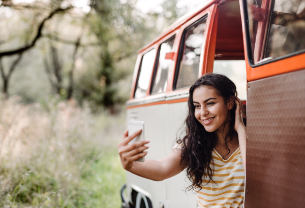 A cheerful young girl with smartphone by a car on a roadtrip through countryside, taking selfie.