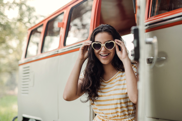 A cheerful young girl with sunglasses standing by a car on a roadtrip through countryside.