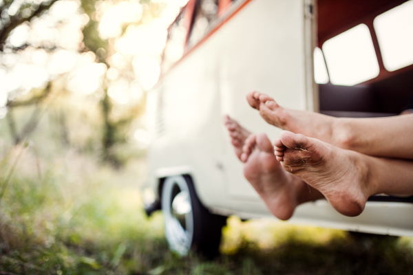 A group of young friends standing by a retro minivan on a roadtrip through countryside, feet sticking out.