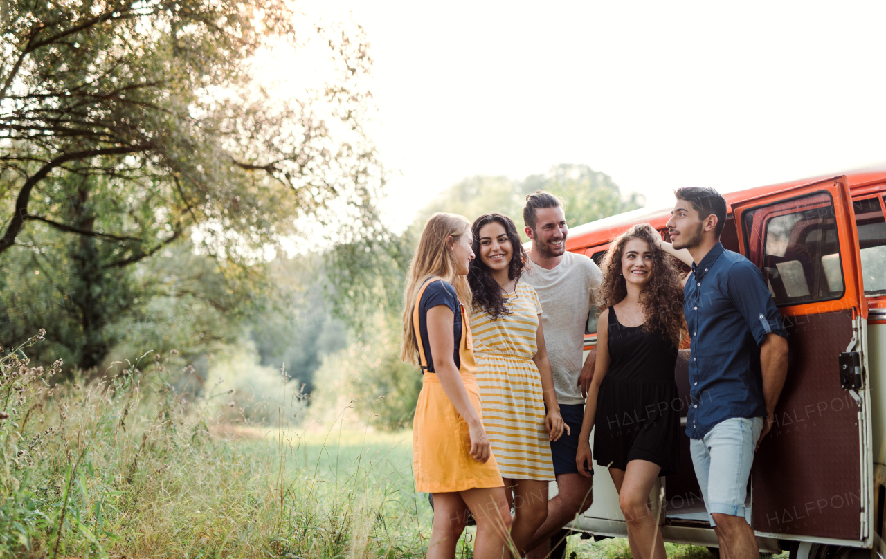 A group of young friends standing by a retro minivan on a roadtrip through countryside.