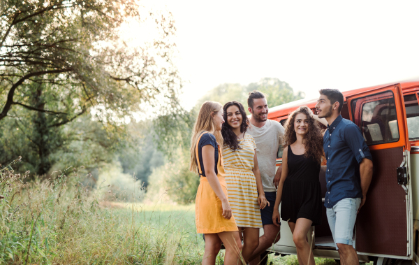 A group of young friends standing by a retro minivan on a roadtrip through countryside.