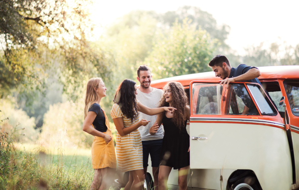 A group of young friends standing by a retro minivan on a roadtrip through countryside.