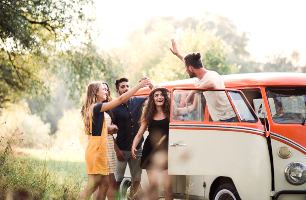 A group of young friends standing by a retro minivan on a roadtrip through countryside.