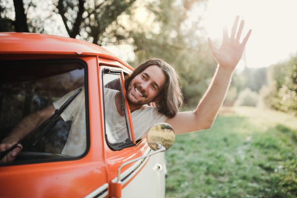 A cheerful young man driver in a car on a roadtrip through countryside, waving at somebody.