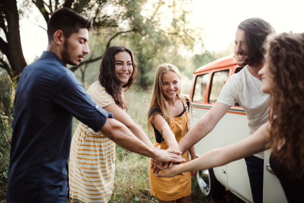 A group of young friends standing by a retro minivan on a roadtrip through countryside, handstack gesture.